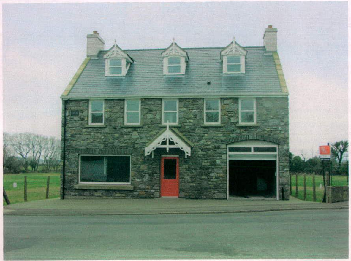Front elevation photograph of a two-story stone building featuring a large ground floor window, a red door with a white canopy, and a garage door.