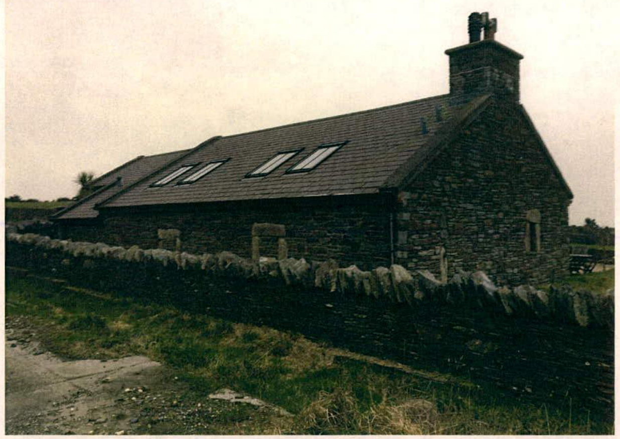 A photograph showing a stone building, likely a converted barn, with a slate roof and skylights behind a dry stone wall.