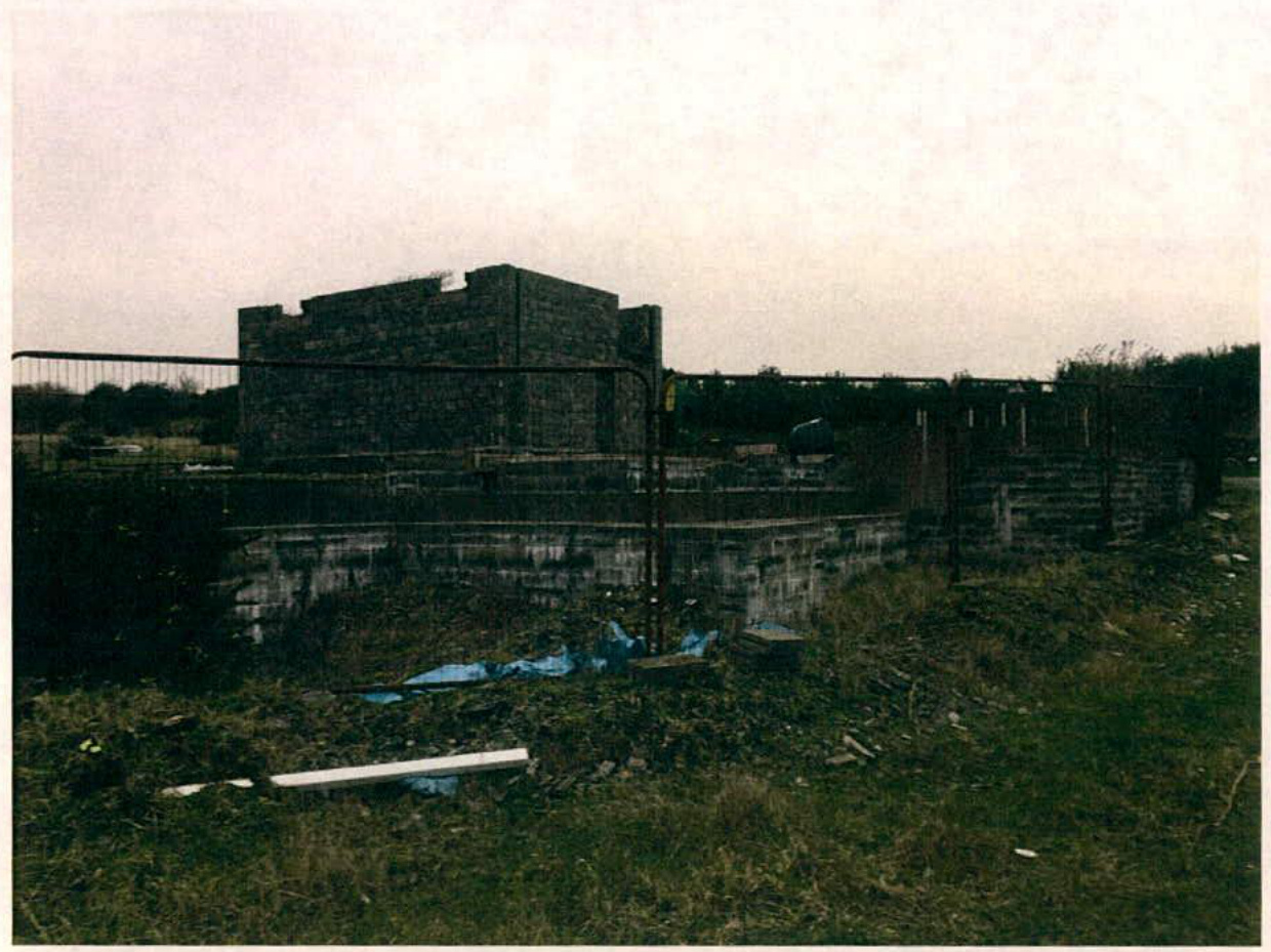 A photograph showing a large stone building structure behind a metal fence and a concrete block retaining wall in a grassy area.