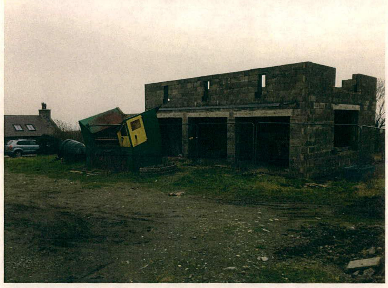 A photograph showing a two-story stone building under construction with large ground floor openings and window cutouts on the upper level. A skip and a neighboring house are visible in the background.