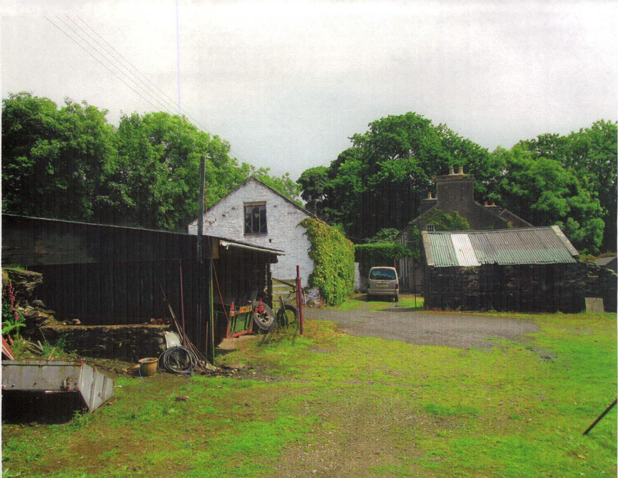 A photograph of a rural property featuring multiple outbuildings, including a white-washed structure and stone barns, set amongst trees with a gravel yard.