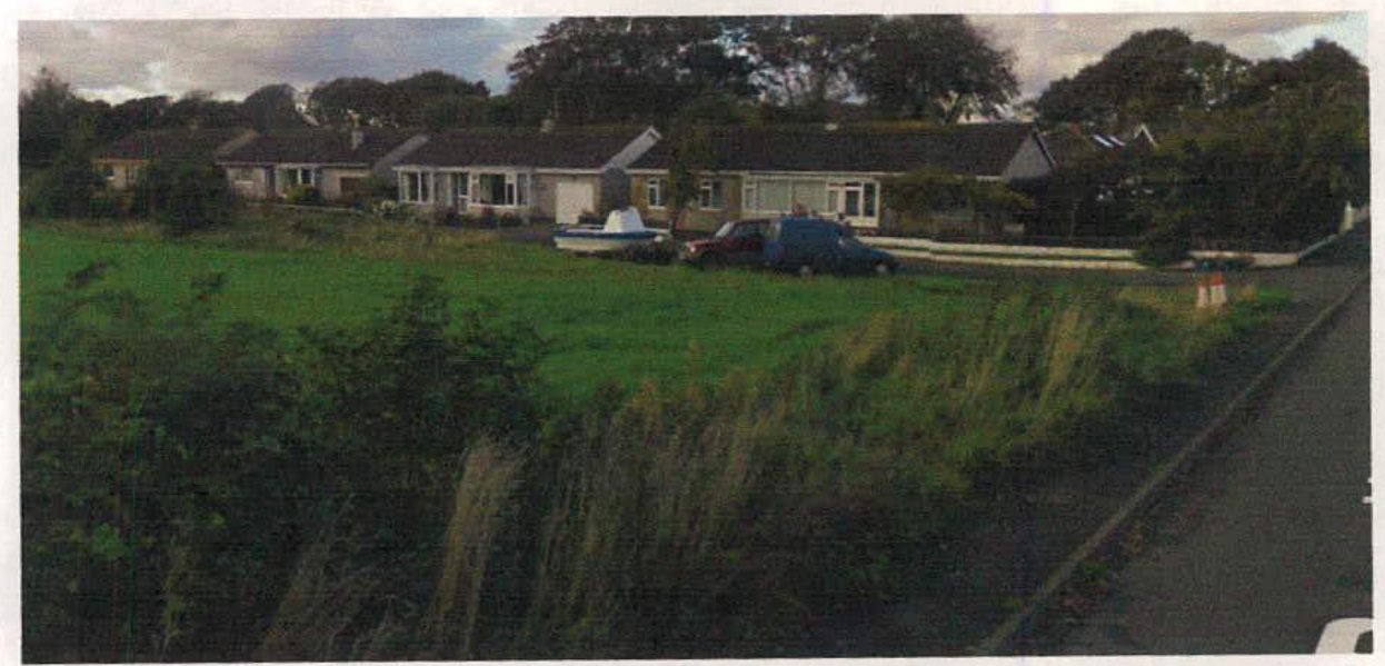 A photograph showing a rural scene with a row of single-story bungalows in the background, a grassy field in the foreground, and a road on the right.