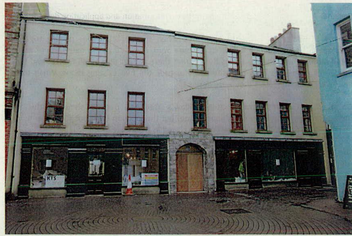Street-level photograph of a three-story building featuring ground-floor retail units and upper-story sash windows, with a boarded-up central archway.