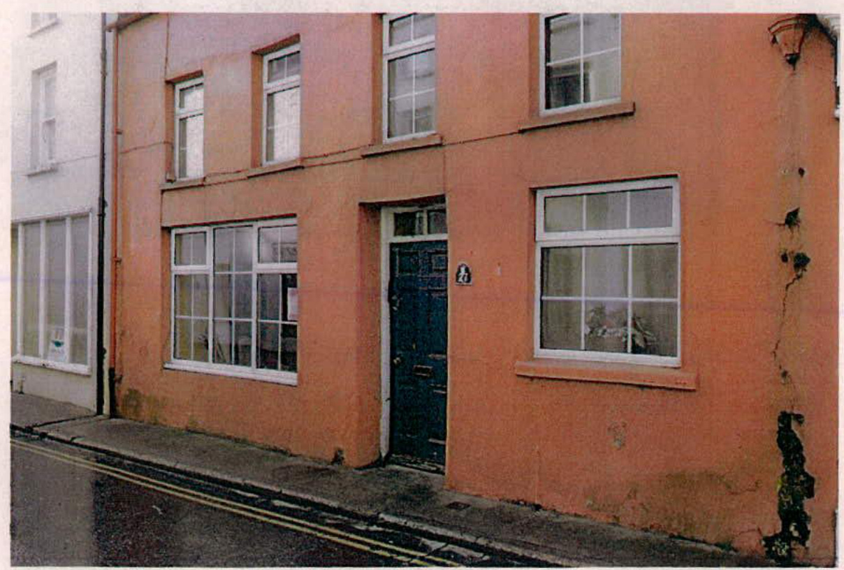 A street-level photograph showing the exterior facade of a two-story salmon-colored building with white-framed windows and a blue door, situated next to a white building.