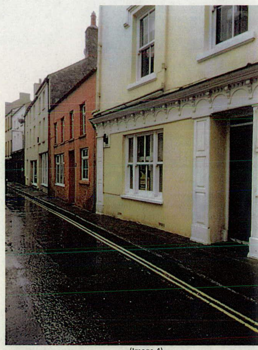 A street-level photograph showing a row of terraced buildings, including a cream-colored building with a large ground-floor window and a reddish-brown building next to it, on a wet street with double yellow lines.