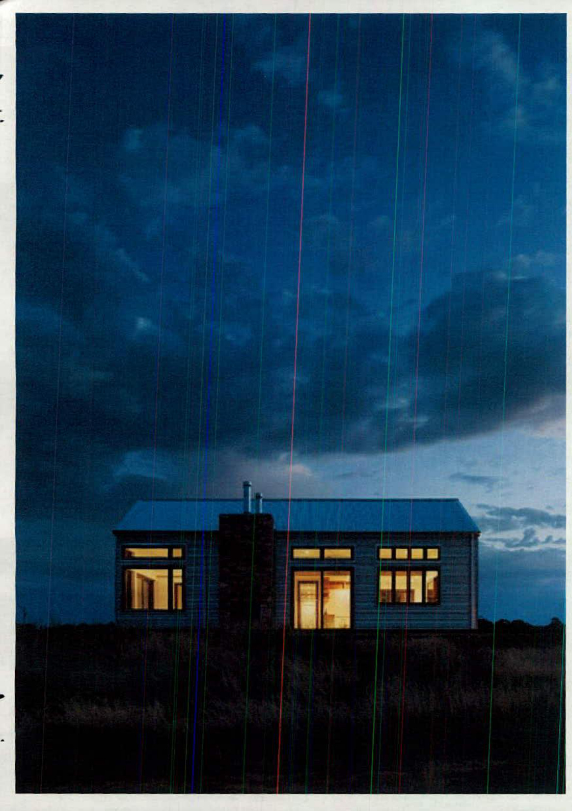 A photograph of a single-story detached house at dusk with warm interior lights glowing through large windows against a dark blue sky.