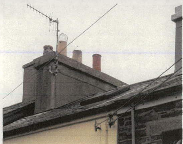 A grainy photograph showing the roof ridge of a building featuring multiple chimneys, slate tiles, and a television aerial.