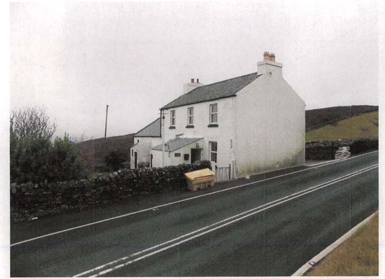 A photograph showing a white, two-story detached house situated alongside a road with a stone wall boundary.