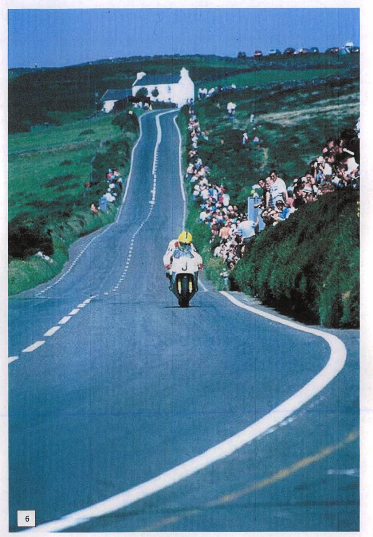 A photograph showing a winding rural road with a motorcyclist riding uphill, lined by crowds of spectators on grassy banks, with a white house visible in the background.