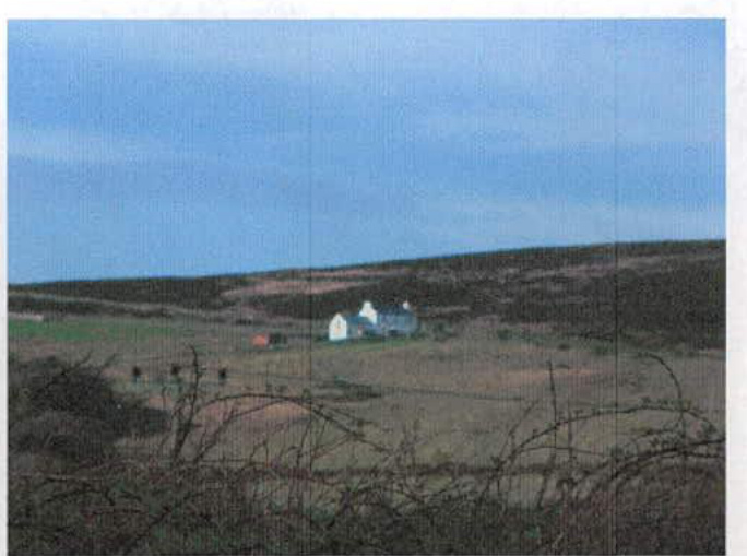 A grainy, distant photograph showing a white building situated in a rural landscape with rolling hills and fields.