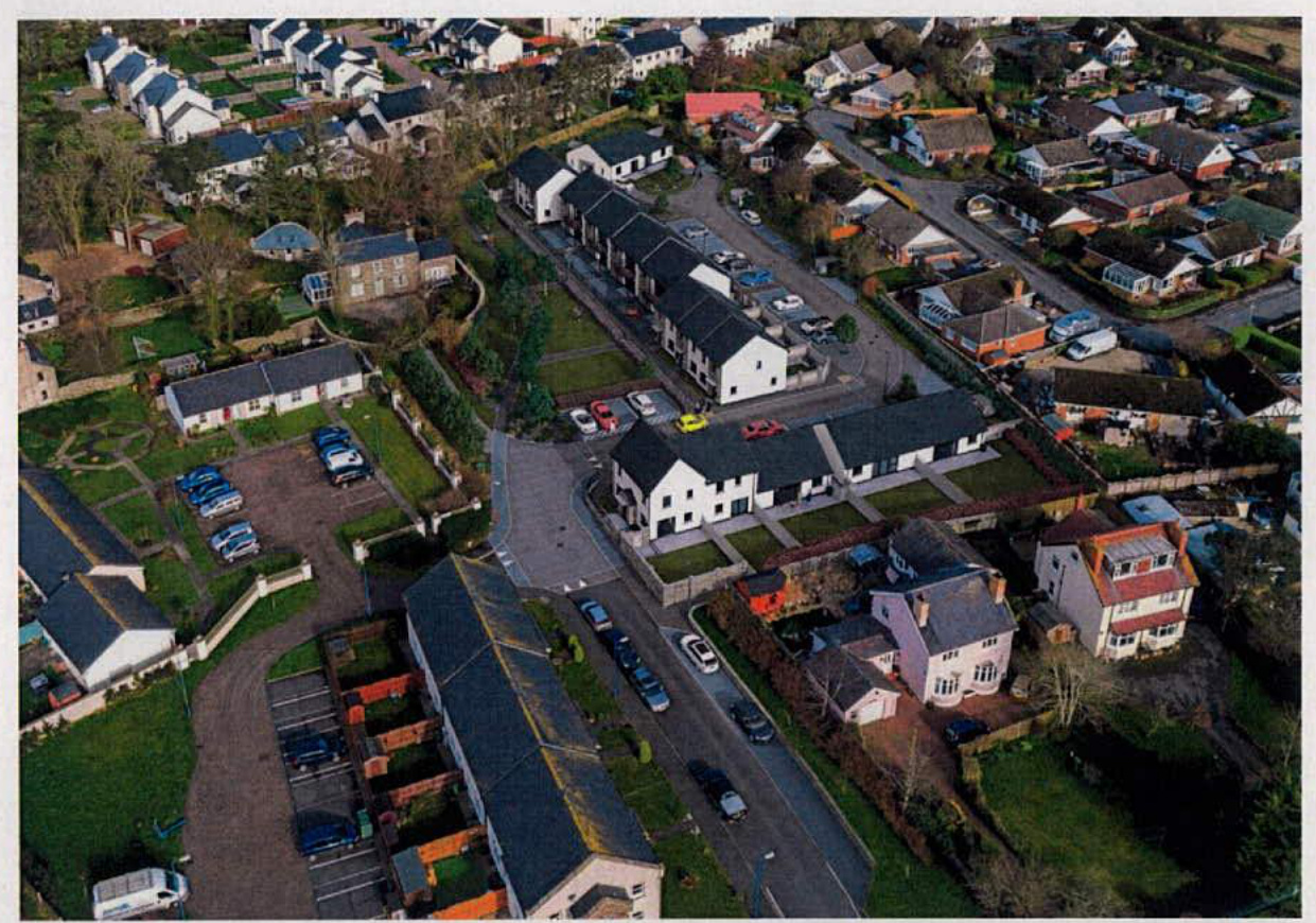 An aerial photograph showing a residential street with a row of new white terraced houses situated among older existing homes. The scene includes visible road access, parking spaces, and surrounding gardens.