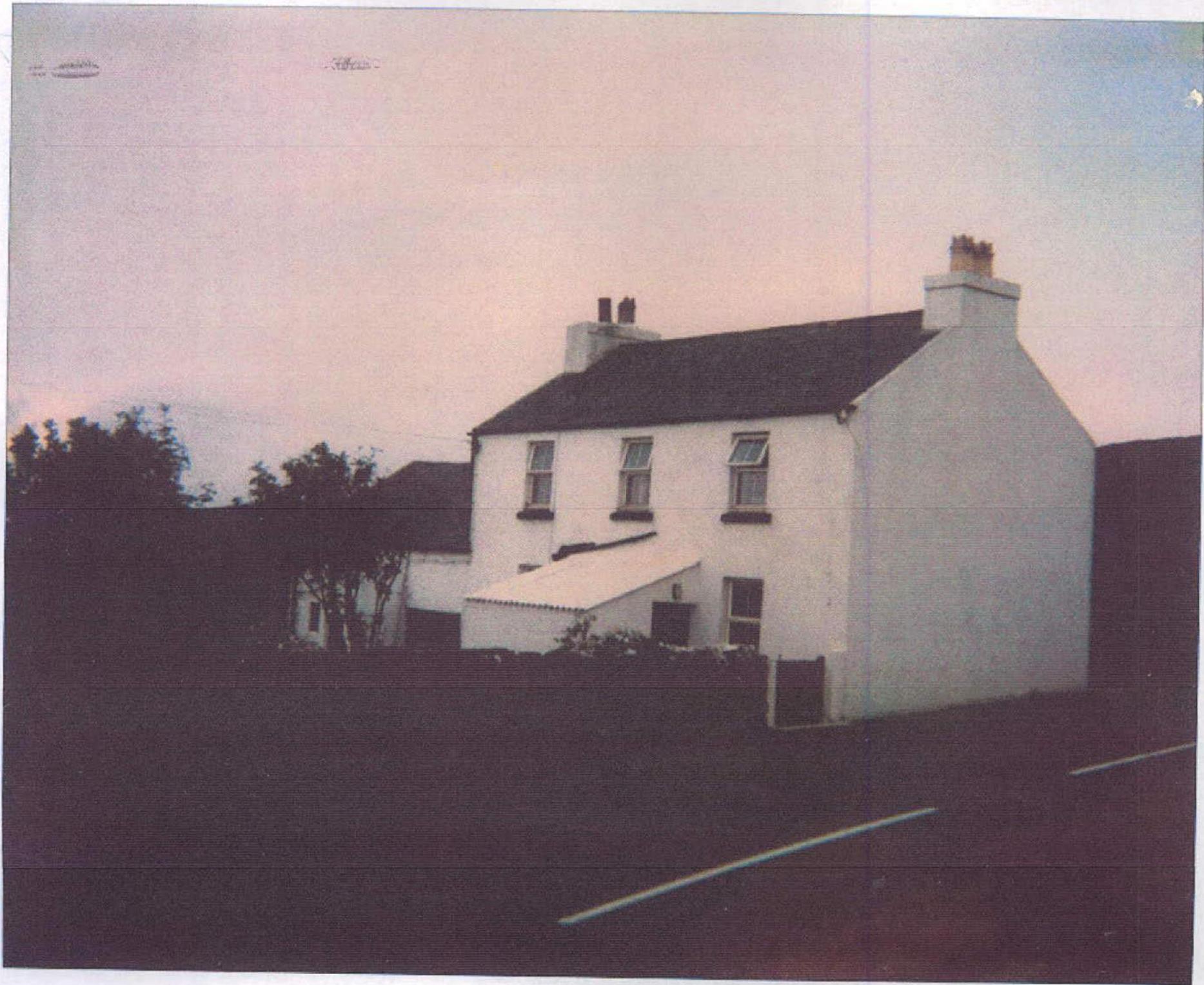 A grainy photograph showing a white, two-story building with a pitched roof and chimneys, featuring a smaller single-story extension attached to the side.