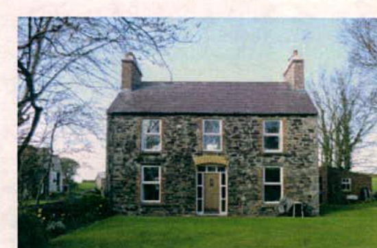 A photograph showing the front elevation of a two-story stone house with a slate roof and white windows.
