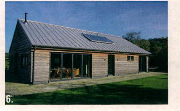 A photograph showing the exterior of a single-story wooden building, likely a converted barn, featuring a metal roof with a solar panel and large glass windows.