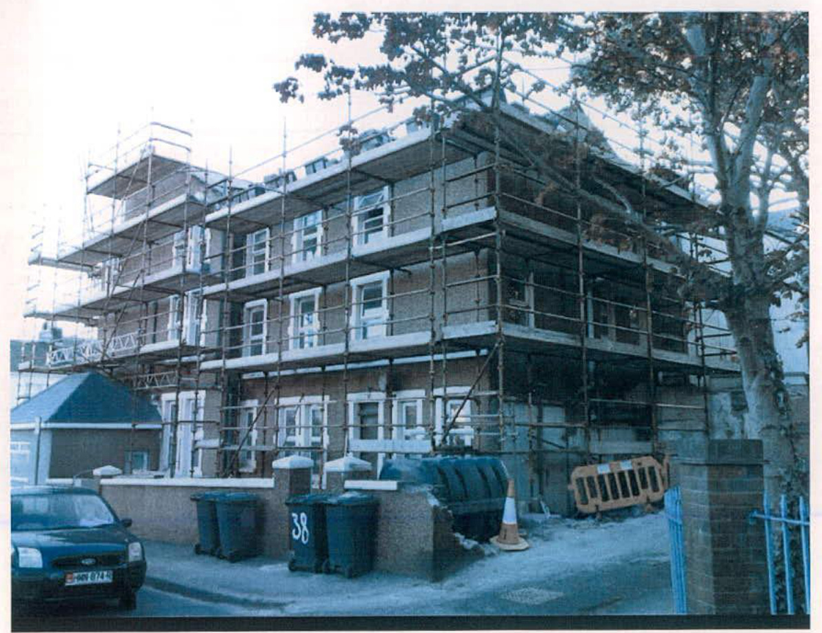 A photograph of a large multi-story brick building covered in scaffolding, likely undergoing renovation or conversion, with a car parked in front and wheelie bins near the boundary wall.