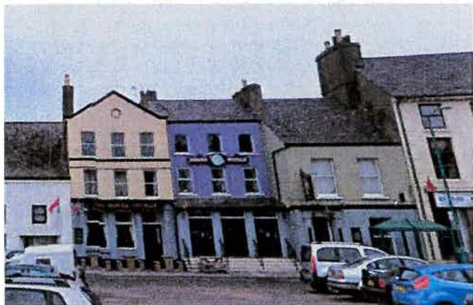 A street-level photograph showing a row of terraced commercial buildings with varied facades, including a yellow gable-fronted building and a purple building, with cars parked in the foreground.