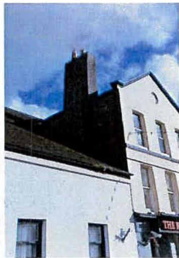 A photograph showing the upper facade of a white building with a prominent dark chimney stack against a blue sky.