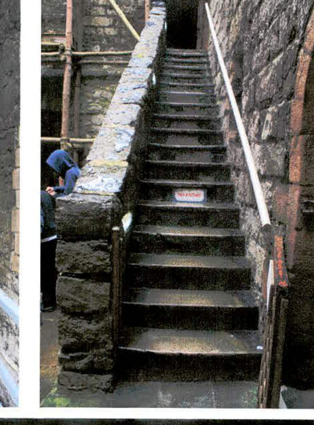 A photograph showing a stone staircase with a white handrail leading up between stone walls, with workers visible on the left side near scaffolding.