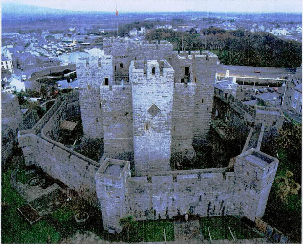 An aerial photograph showing a large medieval stone castle with towers and curtain walls, situated near a coastal town and harbor.