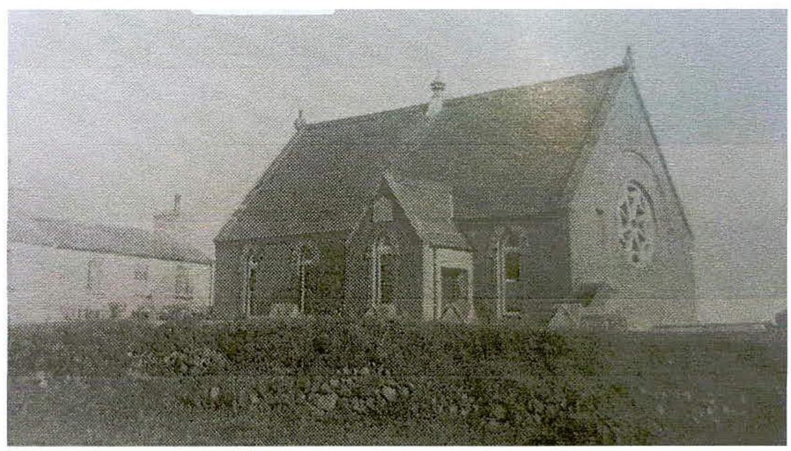 A grainy, monochrome photograph showing the exterior of a small chapel with a pitched roof and a circular window on the gable end.
