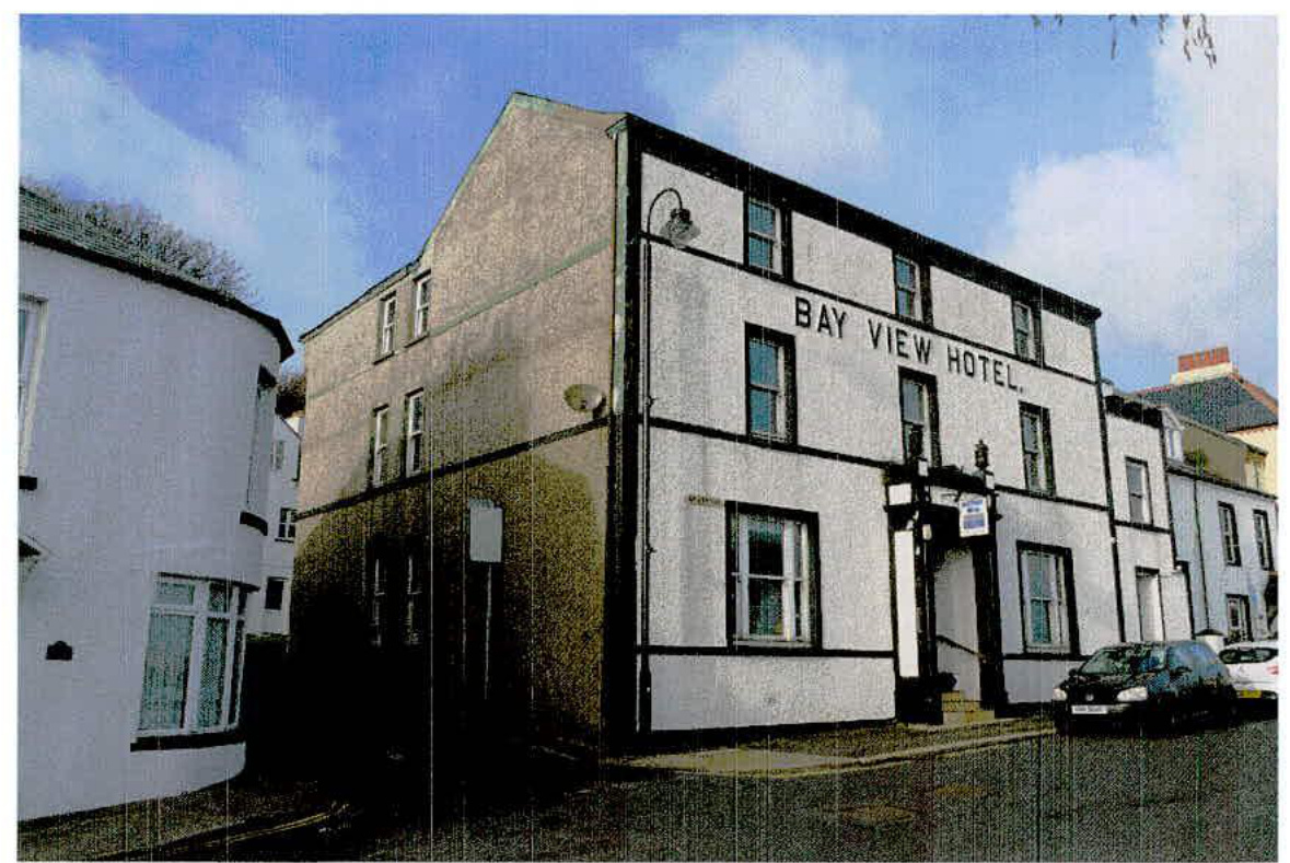A photograph showing the exterior of the existing 'Bay View Hotel', a white three-story commercial building on a street corner.