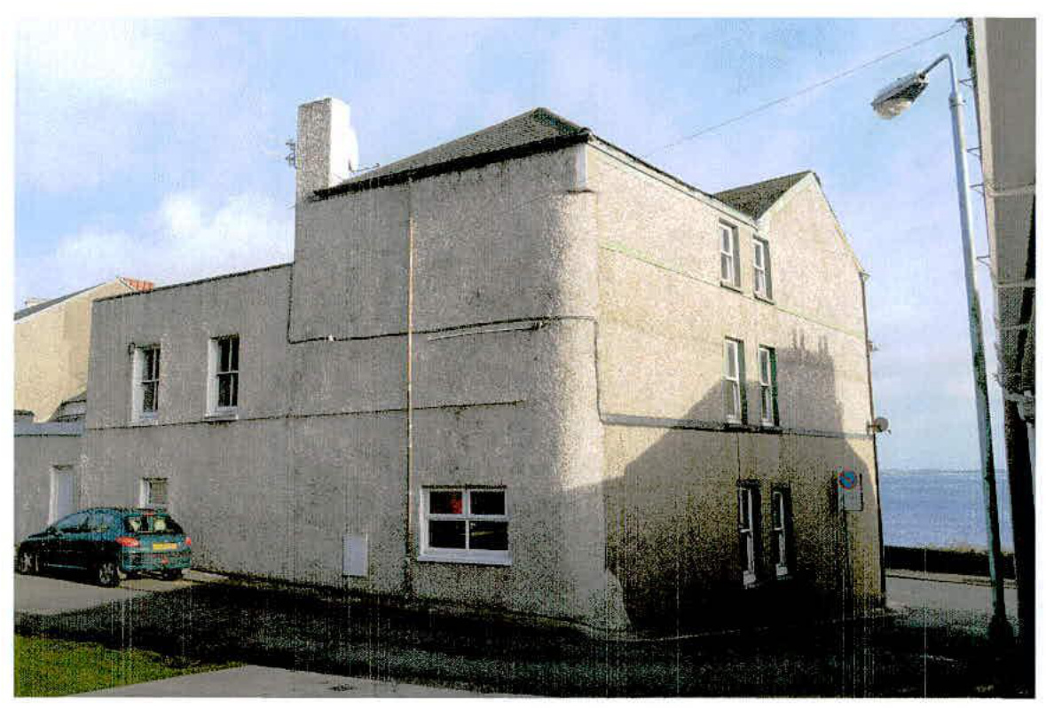 A photograph of the existing two-story building proposed for demolition, featuring roughcast walls and a car parked nearby with the sea visible in the background.