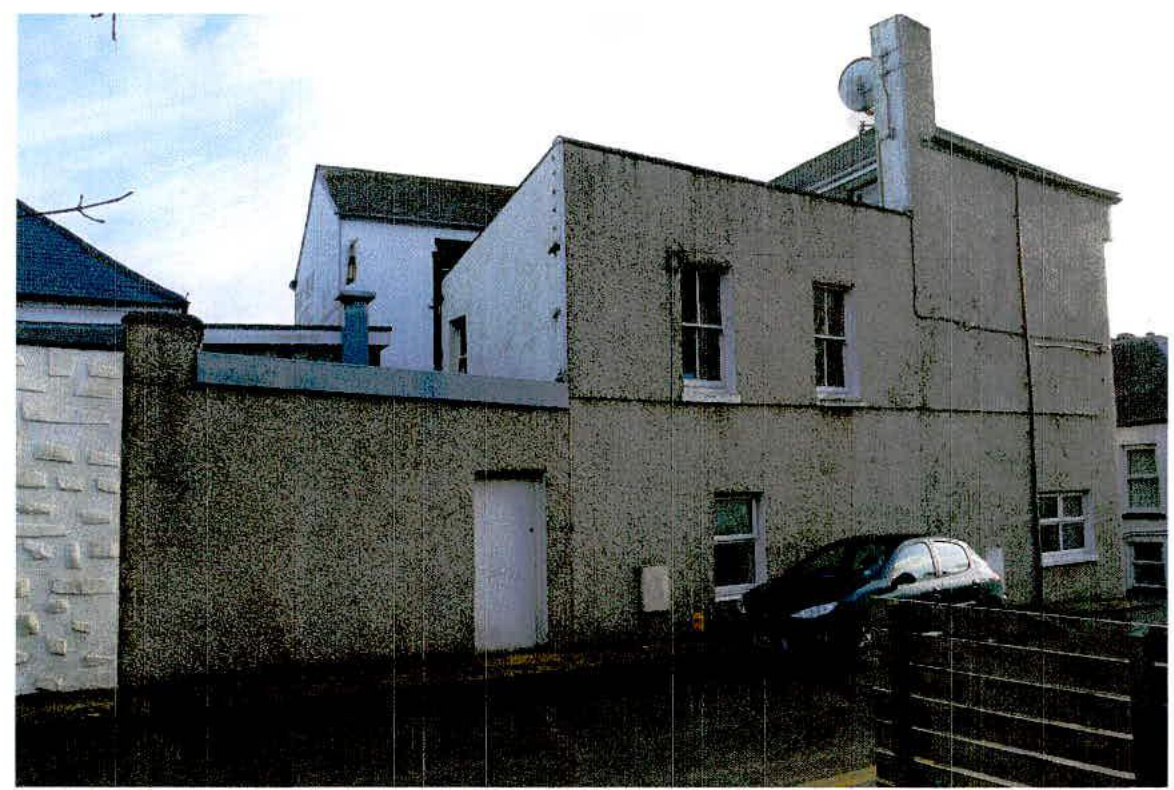 A photograph showing the exterior of a two-story rendered building with a car parked in front, likely the existing structure to be demolished.