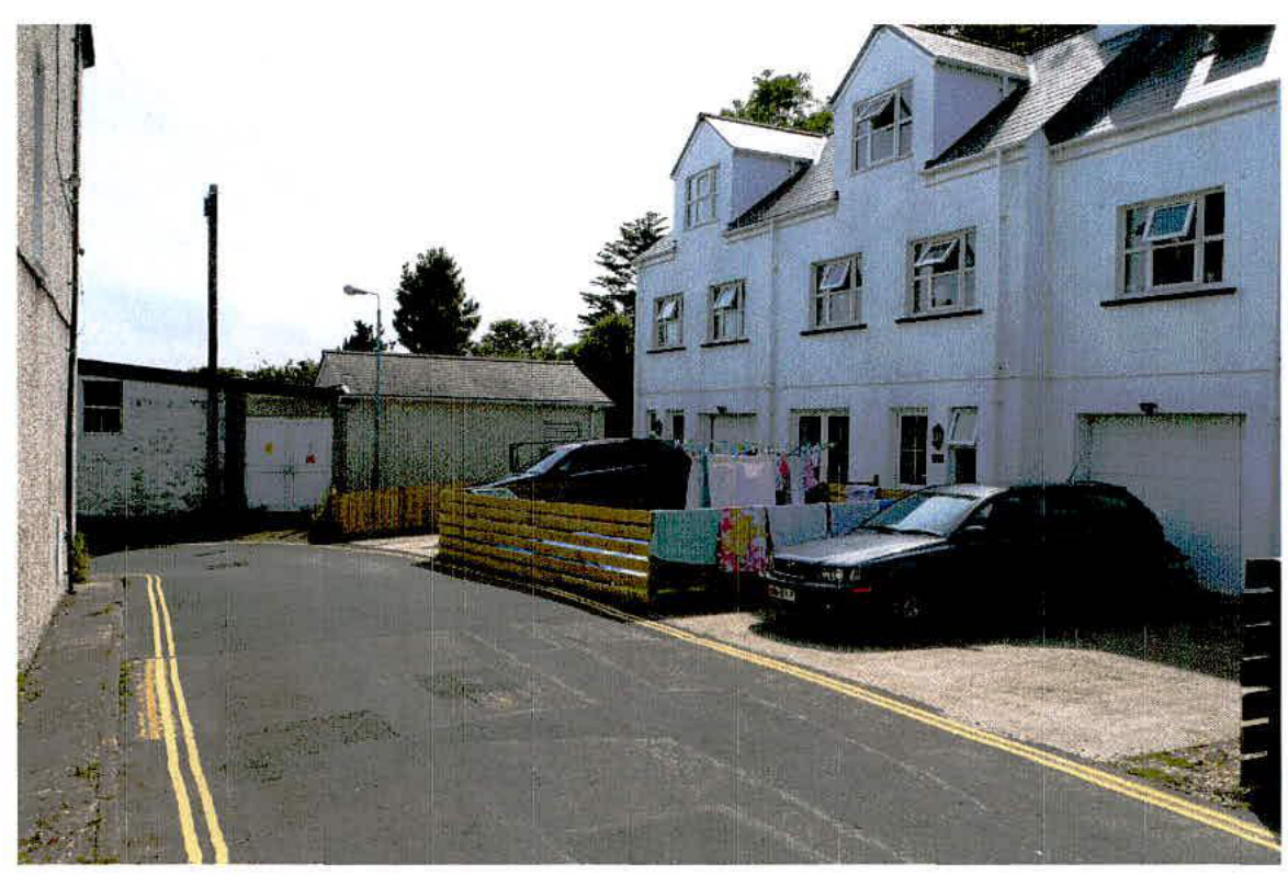 A street-level photograph showing a white residential building with a garage and parked cars, alongside a smaller outbuilding and wooden fencing.