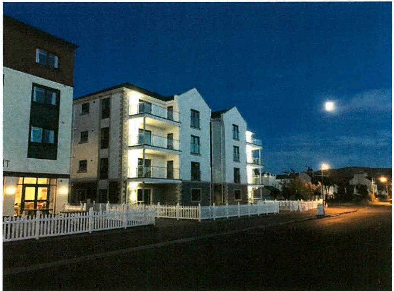 A night-time photograph showing a modern multi-storey residential apartment block with balconies and white cladding, situated behind a white picket fence.