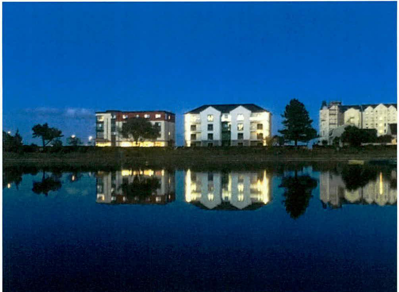 A dusk photograph showing a row of illuminated multi-story residential buildings reflected in a body of water.