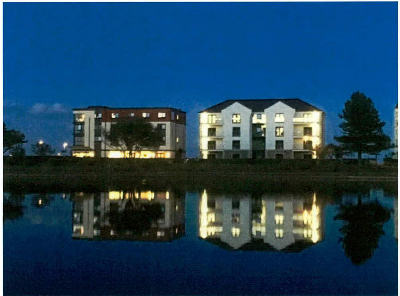 A night-time photograph showing illuminated multi-story residential apartment blocks reflected in a body of water.