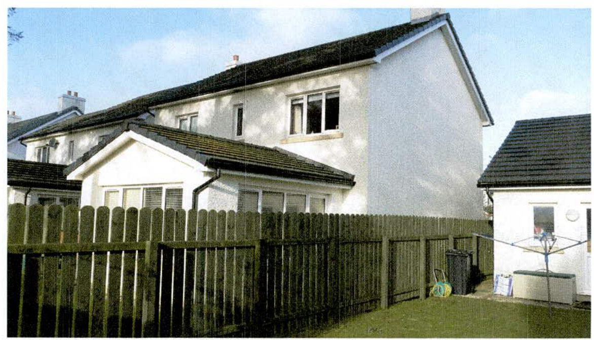 A photograph showing the rear elevation of a white, two-story detached house with a pitched roof and a wooden fence in the foreground.