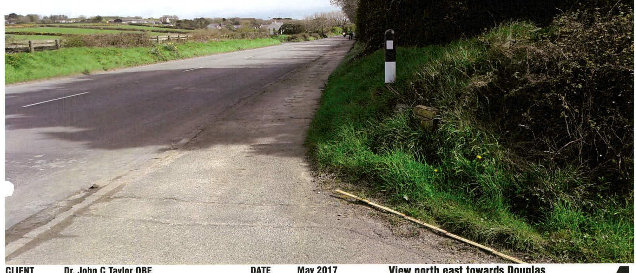 A photograph showing a rural road with a grassy bank and a black and white bollard on the right, captioned as a view north east towards Douglas.