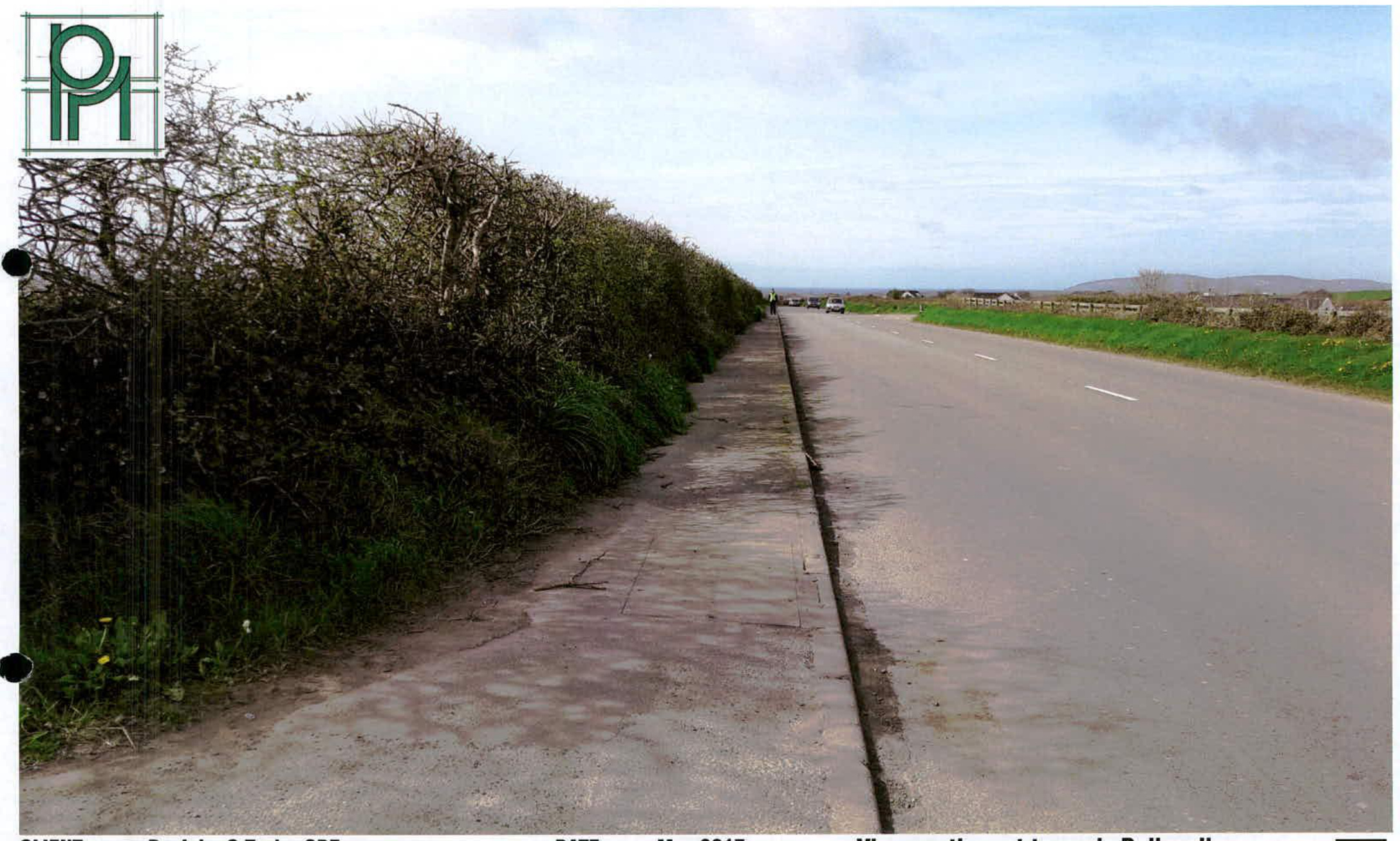 A photograph showing a paved road with a concrete sidewalk running alongside a tall, dense hedge, looking towards distant hills and fields.