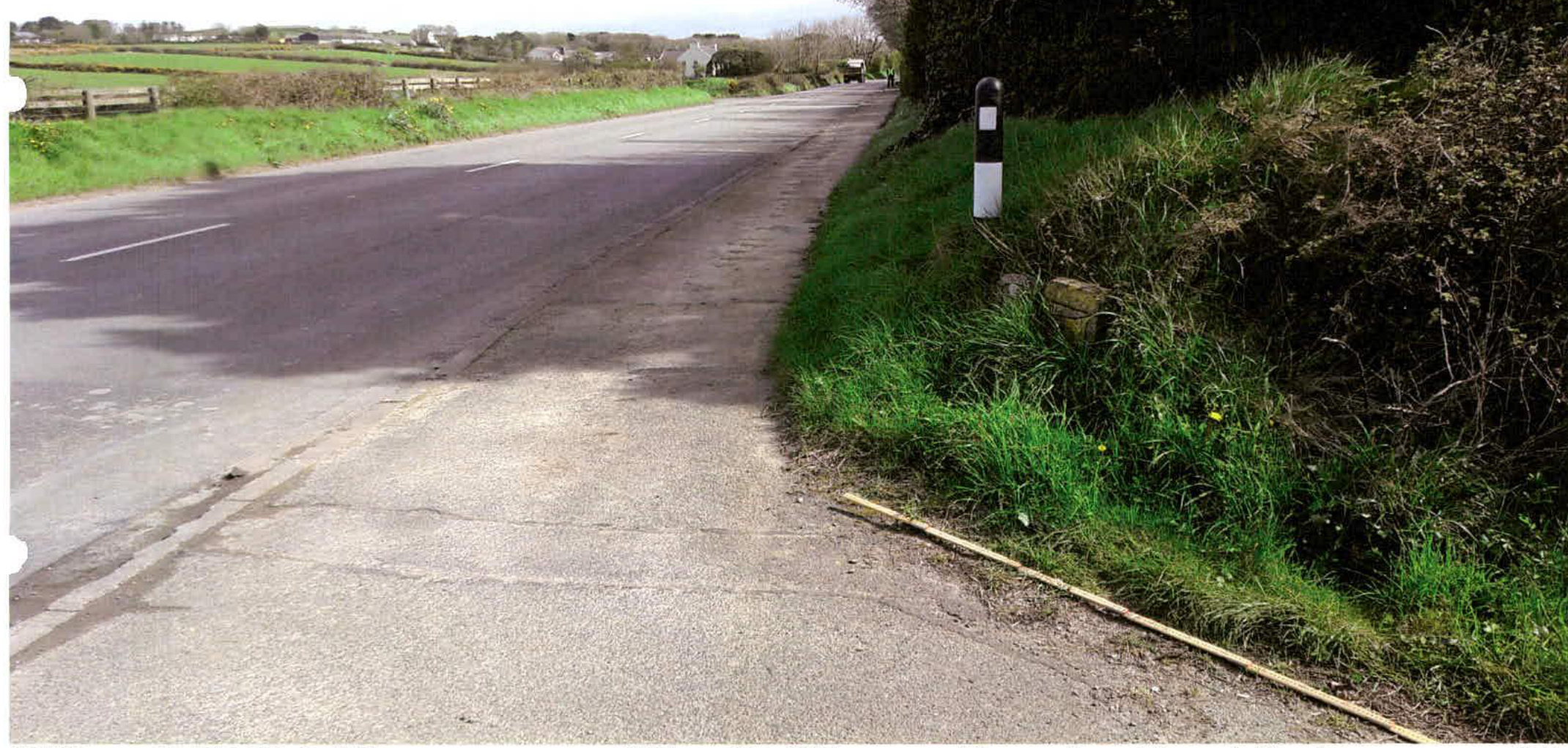 A photograph showing a rural road with a paved access lane or driveway entrance on the right side, marked by a black and white bollard and bordered by a grassy verge and hedge.