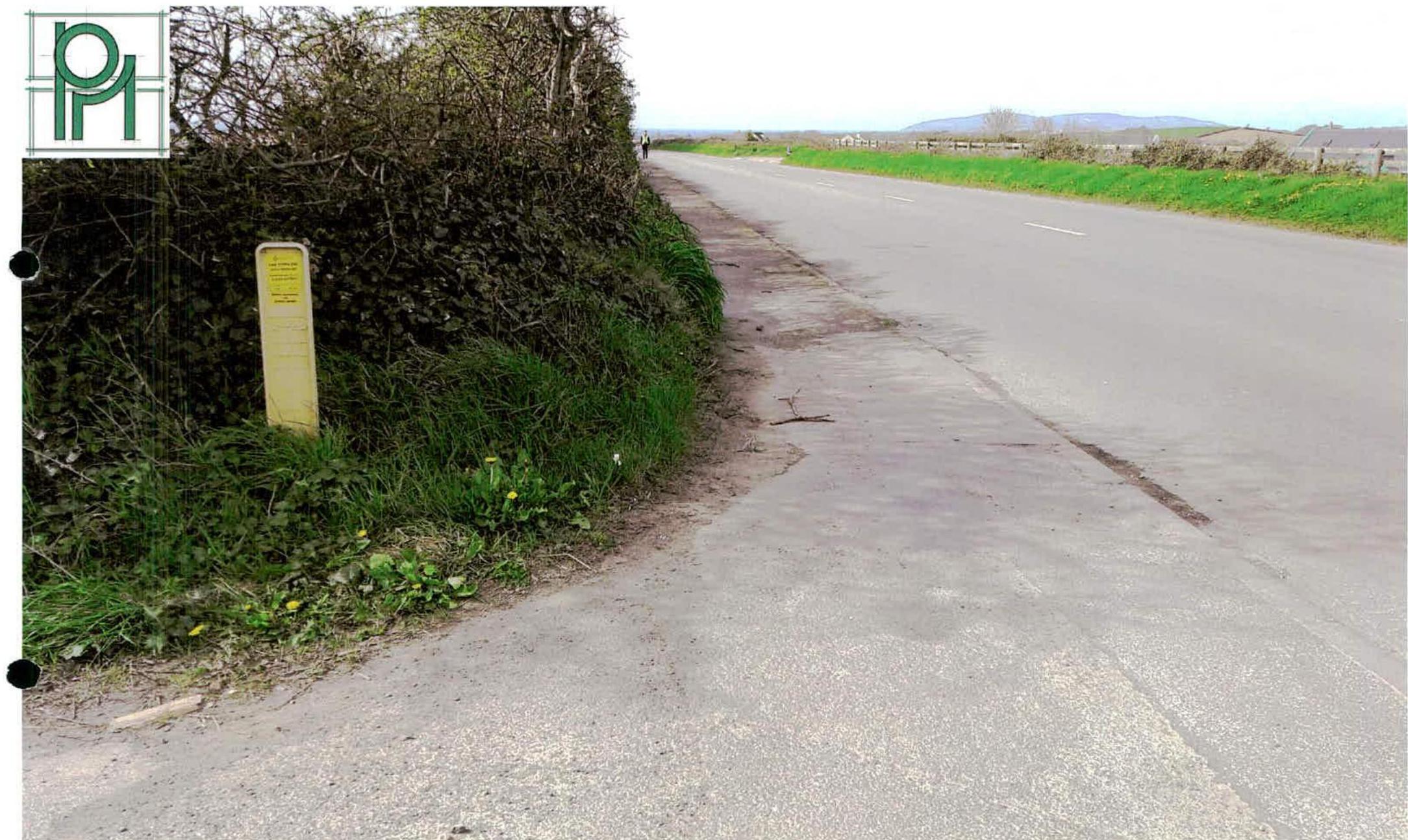 A photograph showing a rural road verge with a dense hedge and a yellow marker post, looking down the lane towards distant hills.