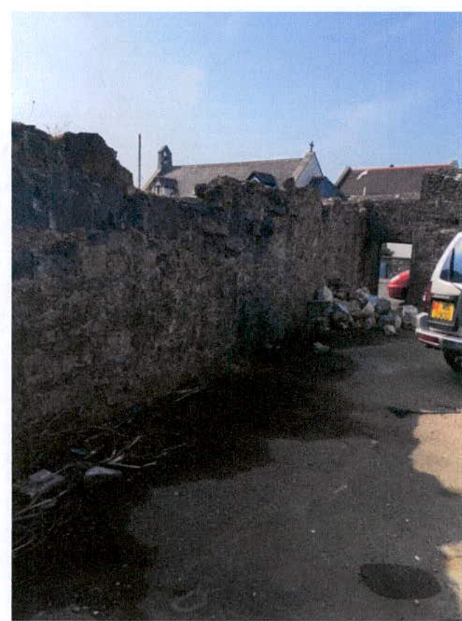 A photograph showing a dilapidated stone wall or ruin with a white van parked nearby and buildings in the background.