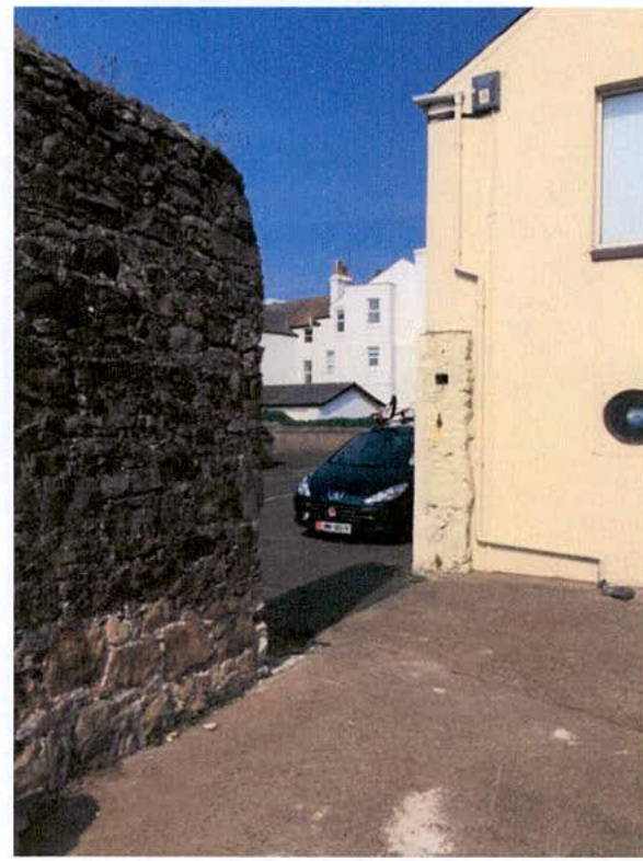 A photograph showing a narrow paved access way between a rough stone wall on the left and a cream-colored building on the right, with a car parked in the background.