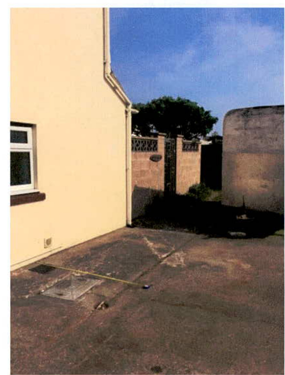 A photograph showing the side elevation of a cream-colored building adjacent to a paved driveway area with a measuring tape on the ground.