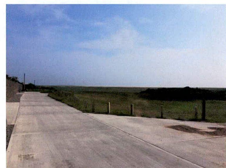 A photograph showing a paved driveway or access road extending towards a flat, grassy landscape with a coastal horizon in the distance. A stone wall is visible on the left and a fence runs along the right side of the...