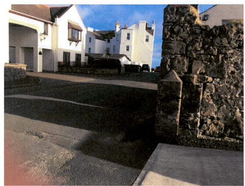 A street-level photograph showing a paved access road and a stone boundary wall in the foreground, with white residential buildings visible in the background.