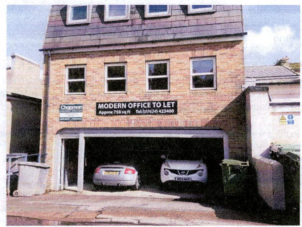 A photograph showing the exterior of a three-story brick building currently used as offices, featuring a ground floor garage with parked cars.
