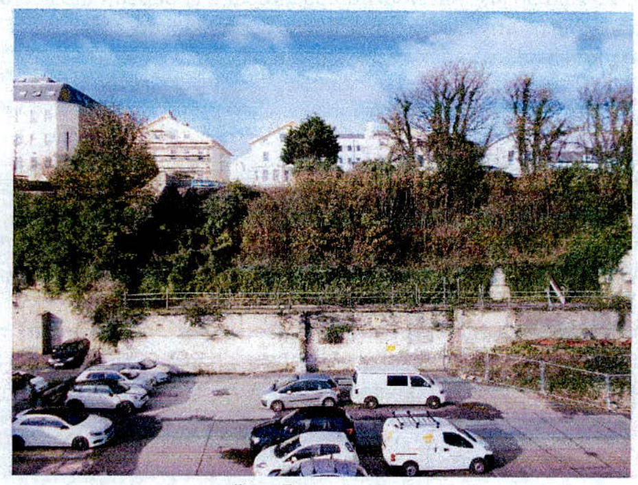 An exterior photograph showing a parking lot in the foreground with a retaining wall and vegetation, behind which stand several large white multi-story buildings.