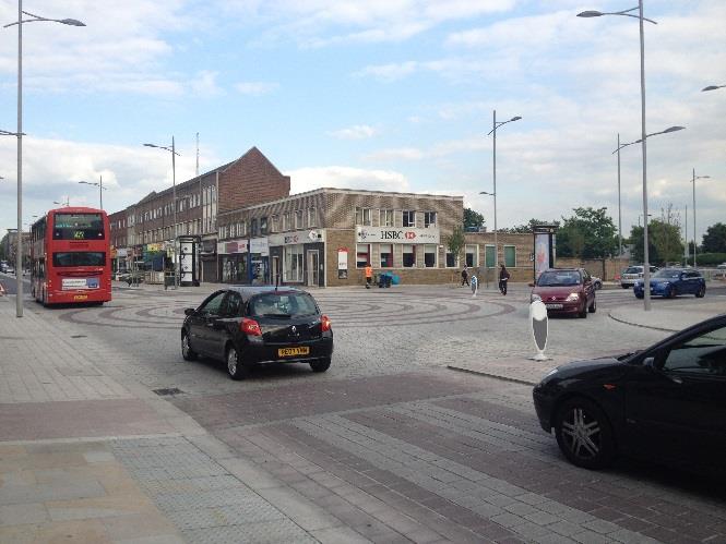 A street-level photograph showing a paved promenade area with a red bus, cars, and commercial buildings including an HSBC branch in the background.