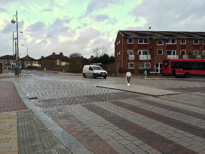 A street-level photograph showing a paved promenade area with patterned brickwork, street furniture, and nearby brick buildings, with a white van and red bus visible.