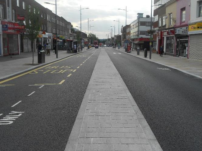 A street-level photograph showing a newly resurfaced promenade with a central paved section and asphalt roads, flanked by commercial buildings and street furniture.