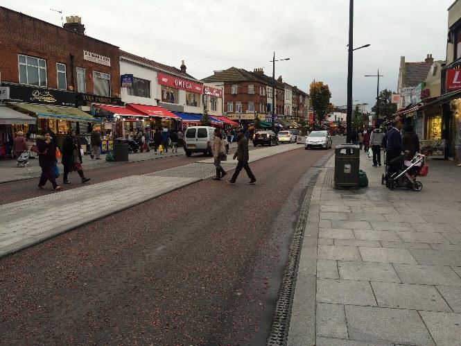 A street-level photograph showing a paved promenade or high street area with pedestrians, shops with awnings, and a visible drainage channel running along the edge.