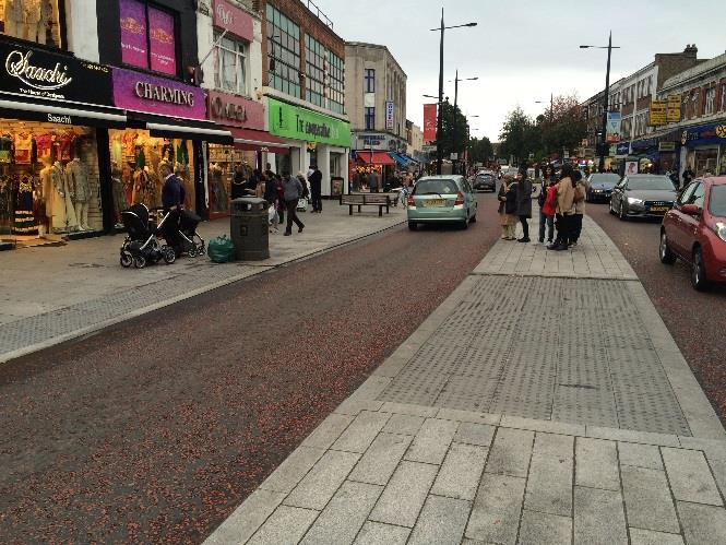 A street-level photograph showing a reconstructed promenade with new paving and road surfacing, featuring shops on the left and pedestrians on the wide footway.