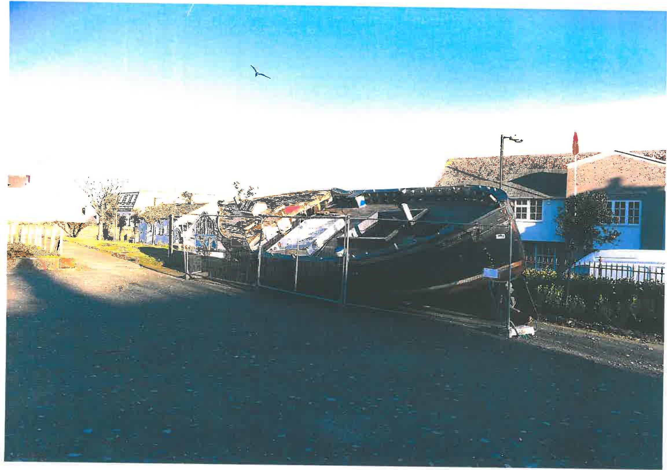 A photograph showing a large boat hull stored on land behind a metal fence, with a road in the foreground and residential buildings in the background.