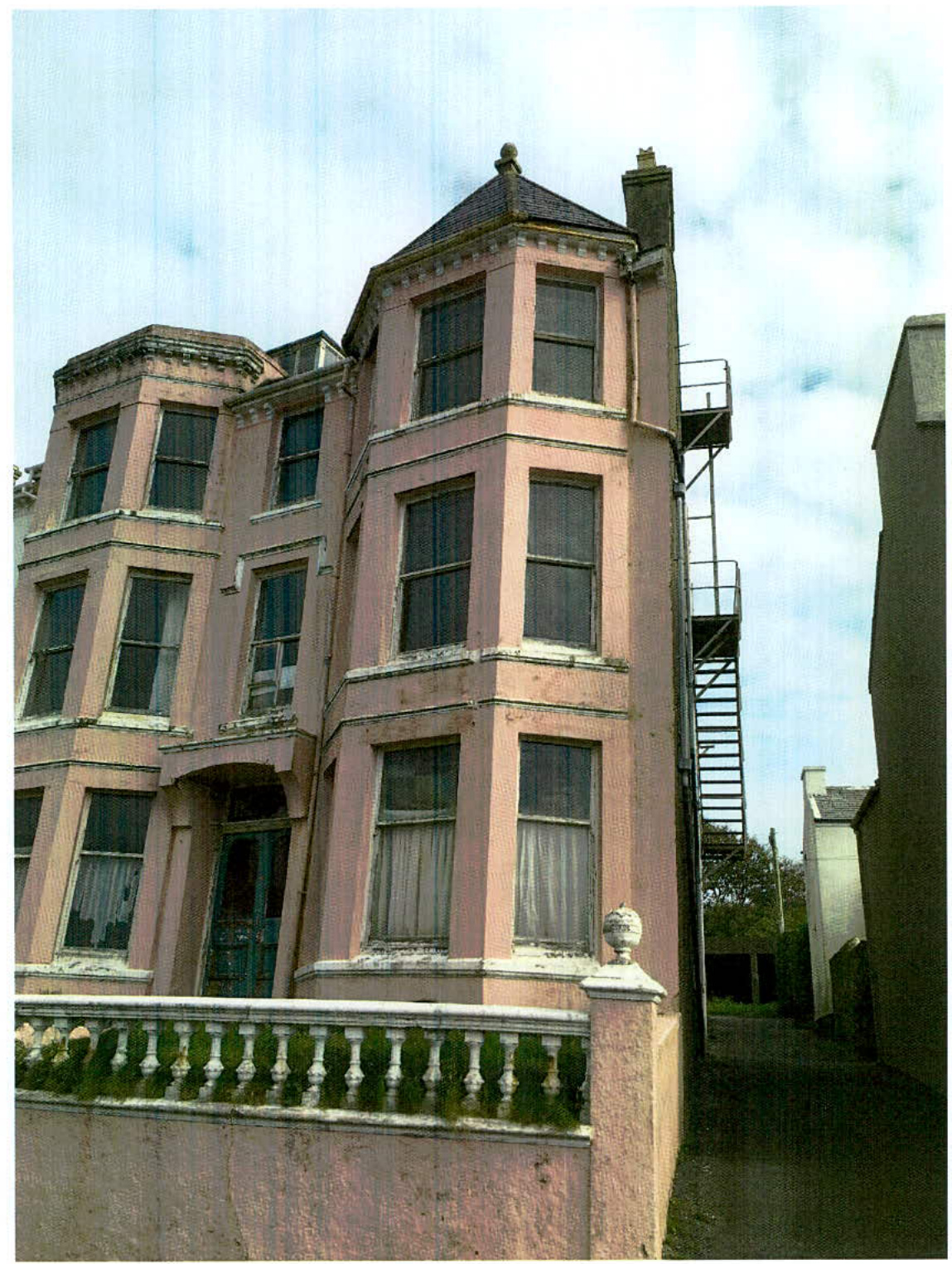 A photograph of a large, pink, multi-story building, likely the former guest house mentioned in the application, featuring a balustrade in the foreground.
